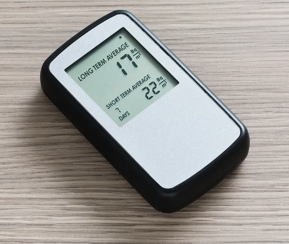 A digital radon detector laying on a wooden surface showing long-term and short-term average indoor gas measurement levels.