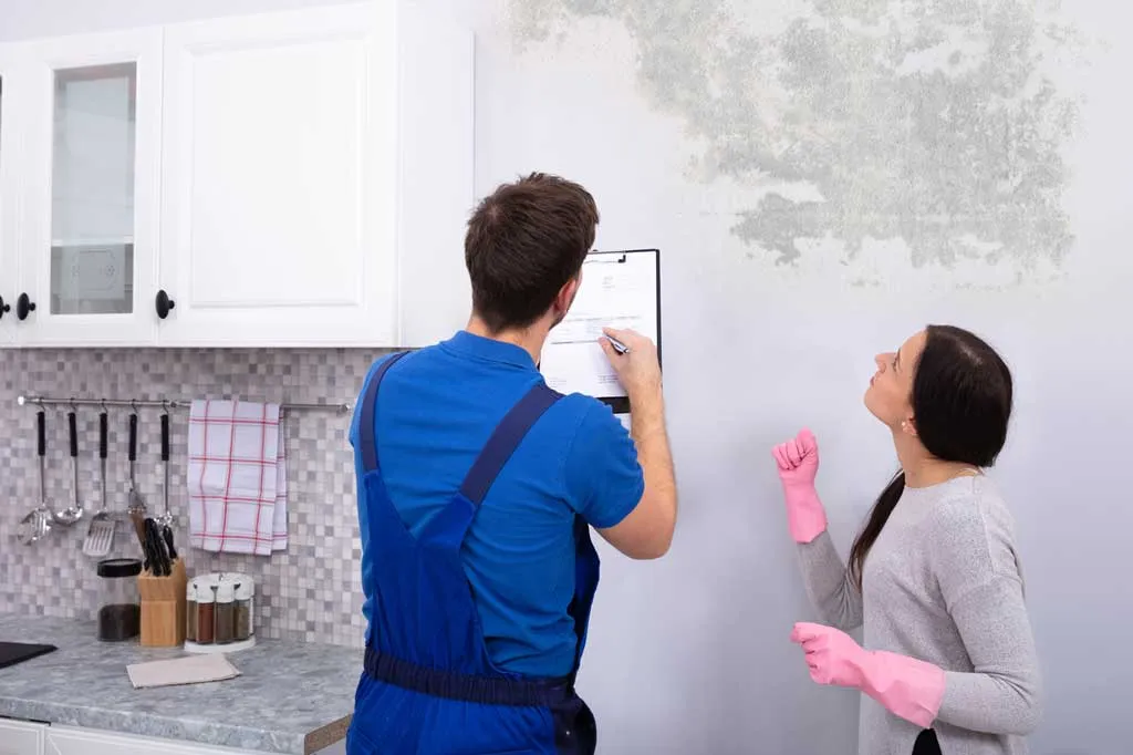 A mold inspector in blue overalls filling out a checklist on a clipboard while a homeowner points out mold growth on a kitchen wall.