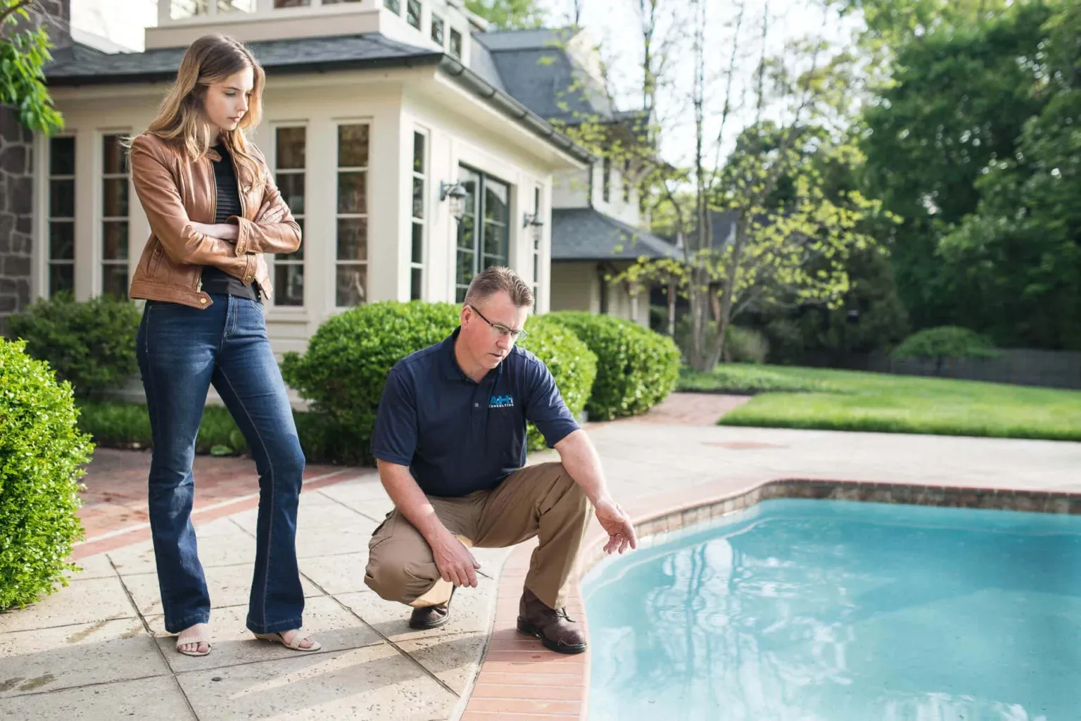 A male property inspector crouching by the edge of a swimming pool, explaining the condition of the water and coping to a female client standing next to him.