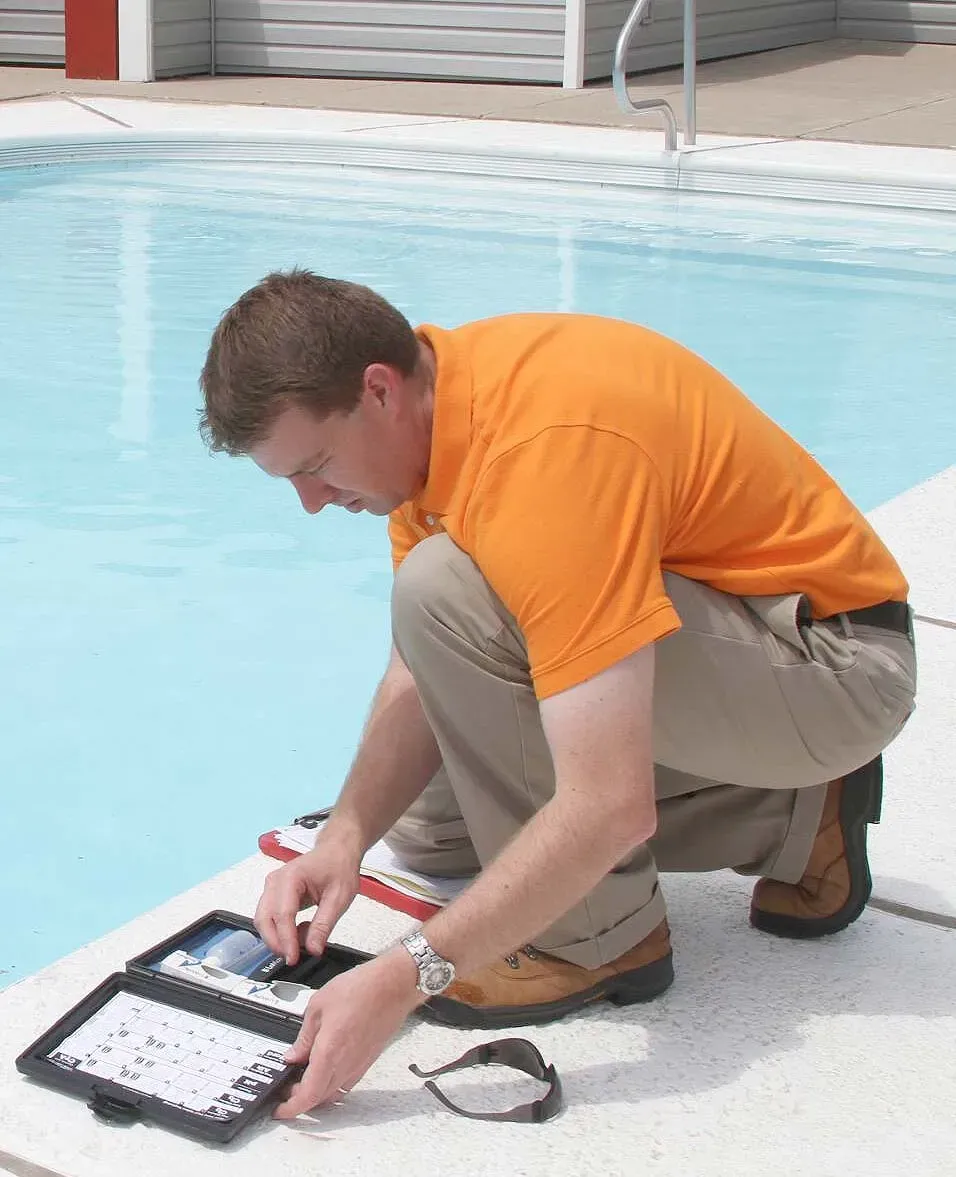 A pool inspector wearing an orange shirt crouching by the edge of an outdoor swimming pool to run a chemical water test.