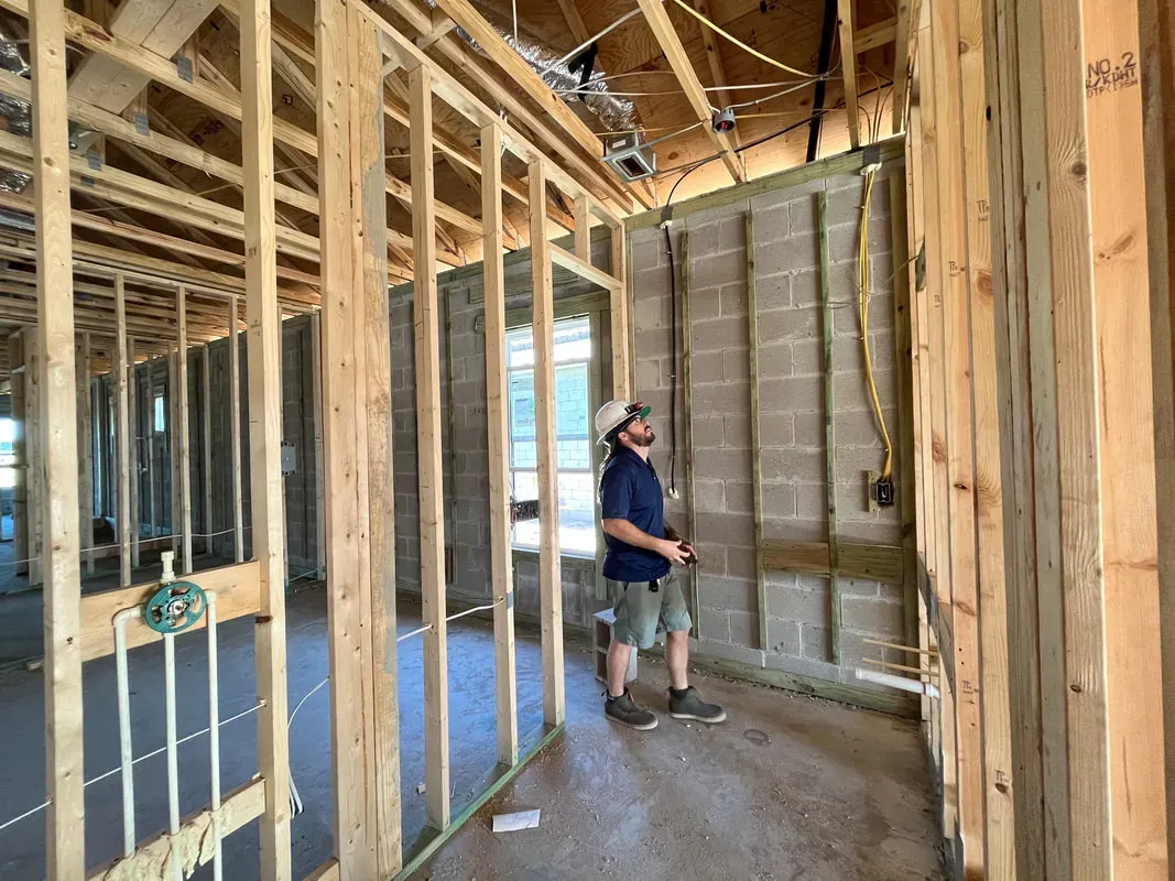 A home inspector wearing a hard hat examining exposed electrical wiring and wood framing inside a house under construction.