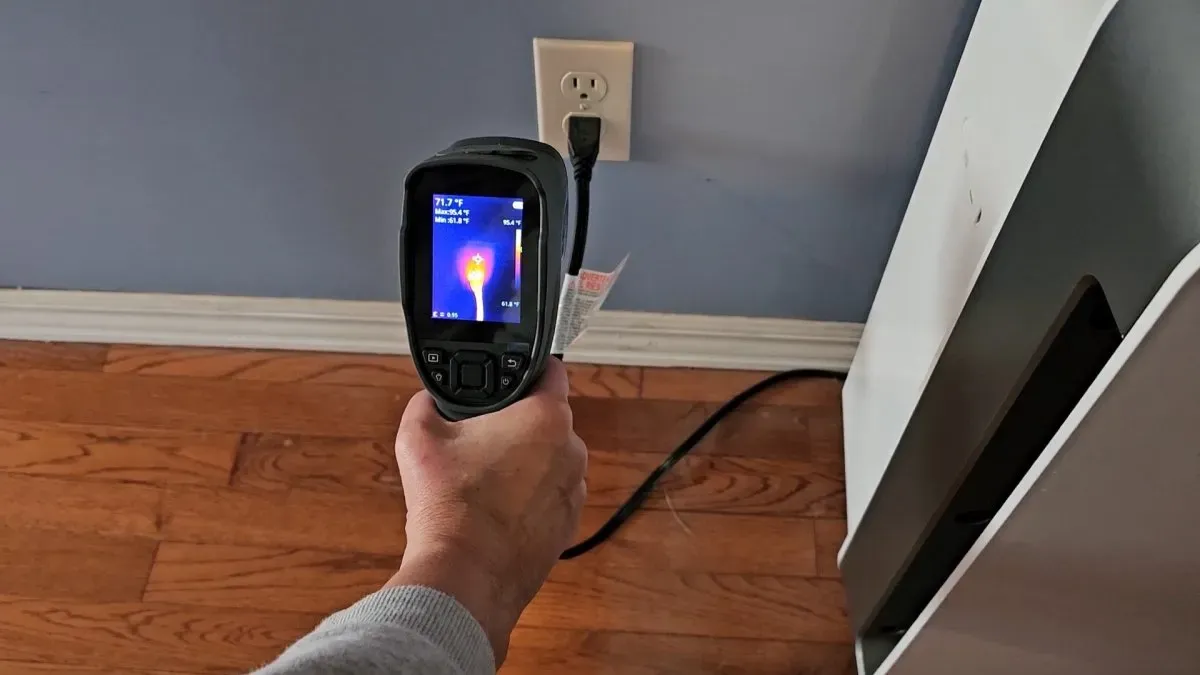 A hand holding an infrared thermal camera showing a bright heat signature from an electrical plug and wall outlet.