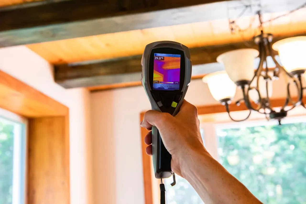 A hand holding an infrared thermal imaging camera showing heat signatures on a residential ceiling and wall joint.